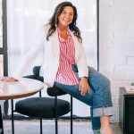 Woman sitting on a chair, smiling, wearing a red and white striped shirt, white blazer, and blue jeans, in a bright room.