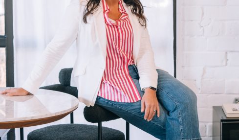 Woman sitting on a chair, smiling, wearing a red and white striped shirt, white blazer, and blue jeans, in a bright room.