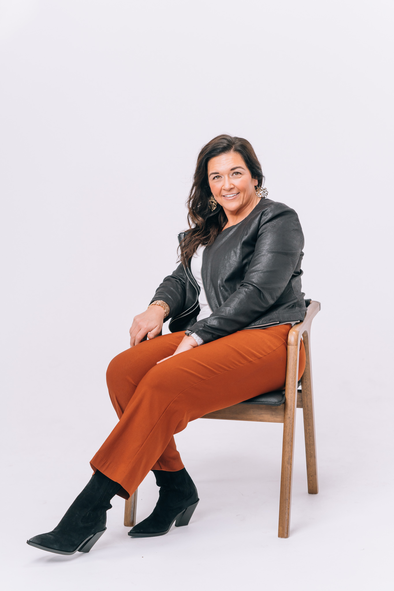 Woman sitting on a chair, smiling, wearing a black leather jacket, white top, orange pants, and black boots.