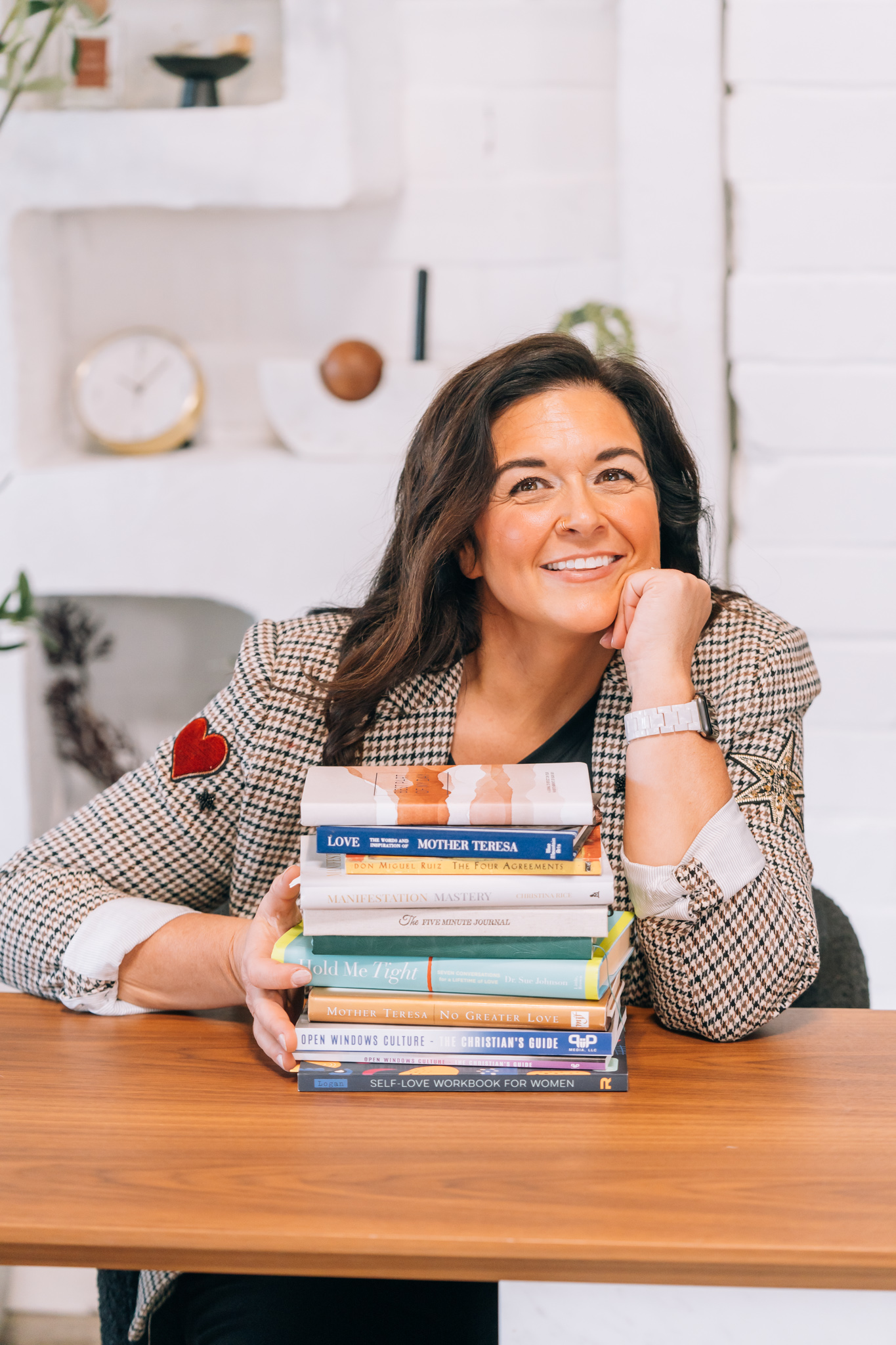 Woman with dark hair smiling, resting her chin on her hand, with a stack of books on a wooden table.