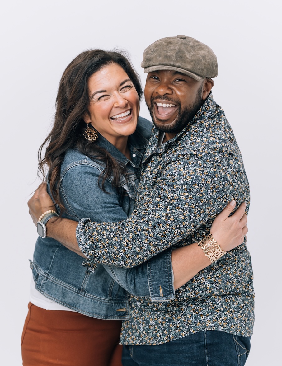 Two people hugging and smiling, woman with long dark hair and man with a cap, against a plain white background.