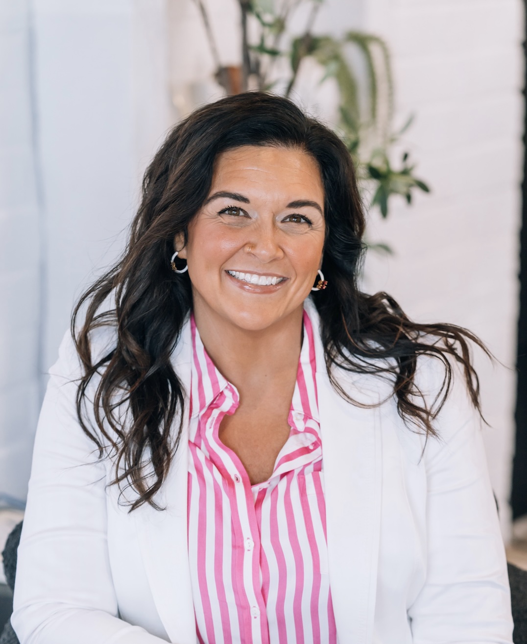 Woman with dark wavy hair smiling, wearing a white blazer and pink striped shirt, in a bright indoor setting.