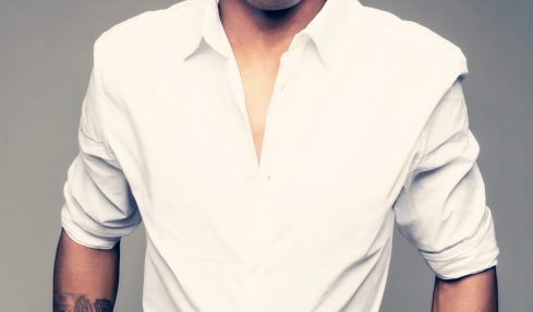 Man with short curly hair wearing a white shirt and dark plaid pants, standing with hands in pockets against a gray background.