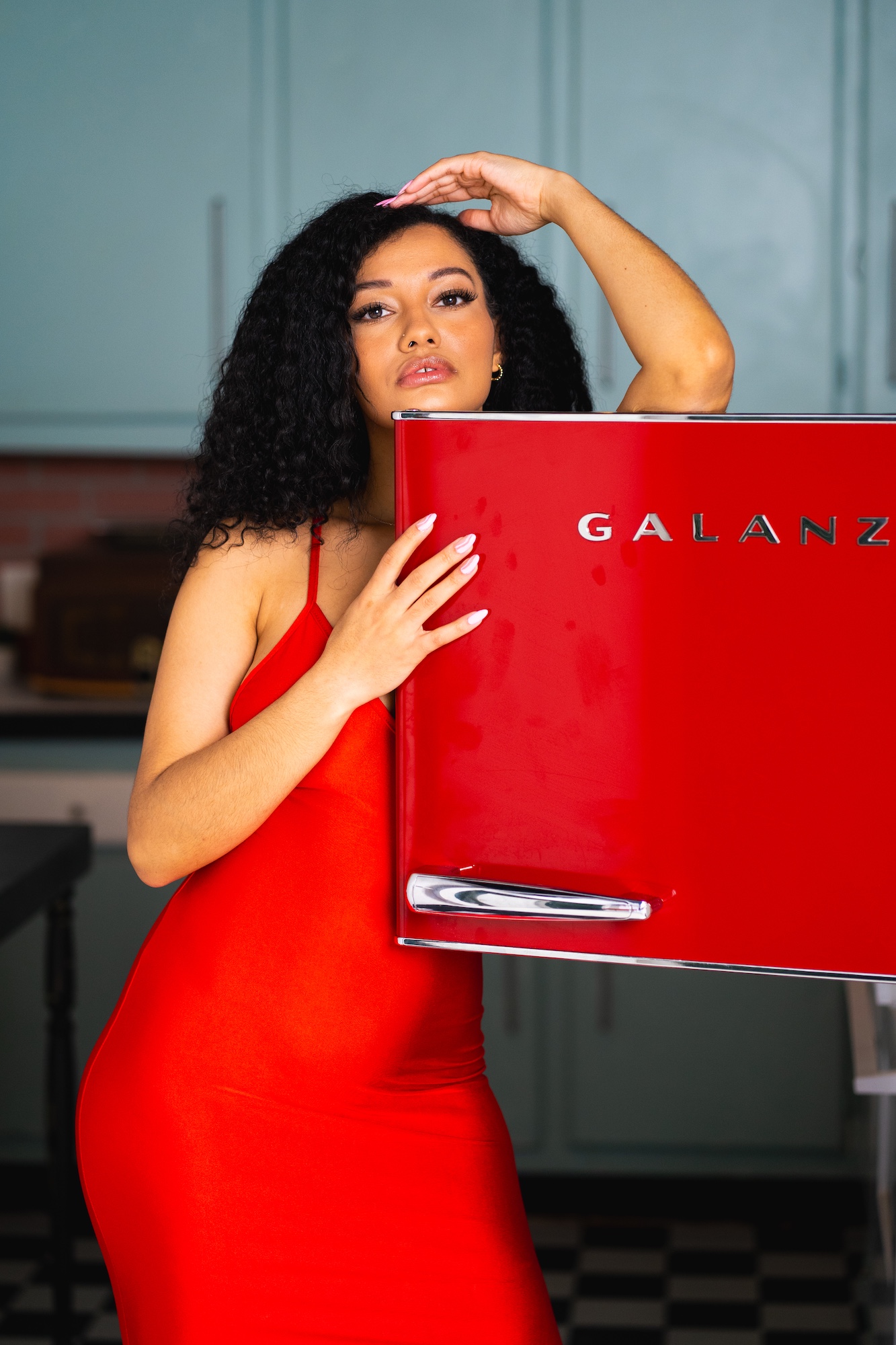 Woman in red dress holding a red Galanz appliance, standing in a room with teal walls and checkered floor.