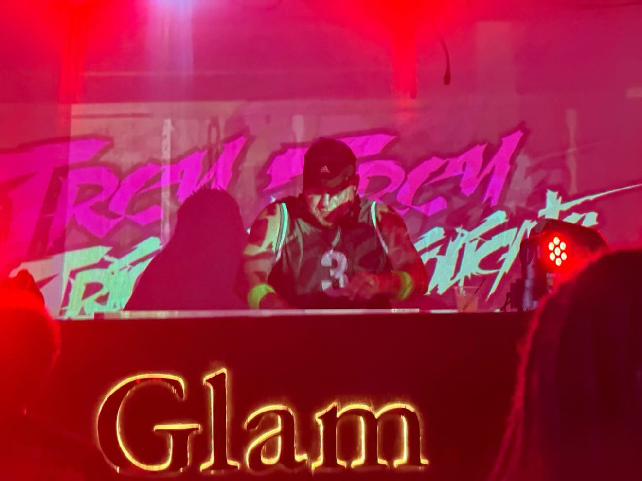 Two people on stage with a red and pink background, one wearing sunglasses and a jacket, behind a sign that says 'Glam'.