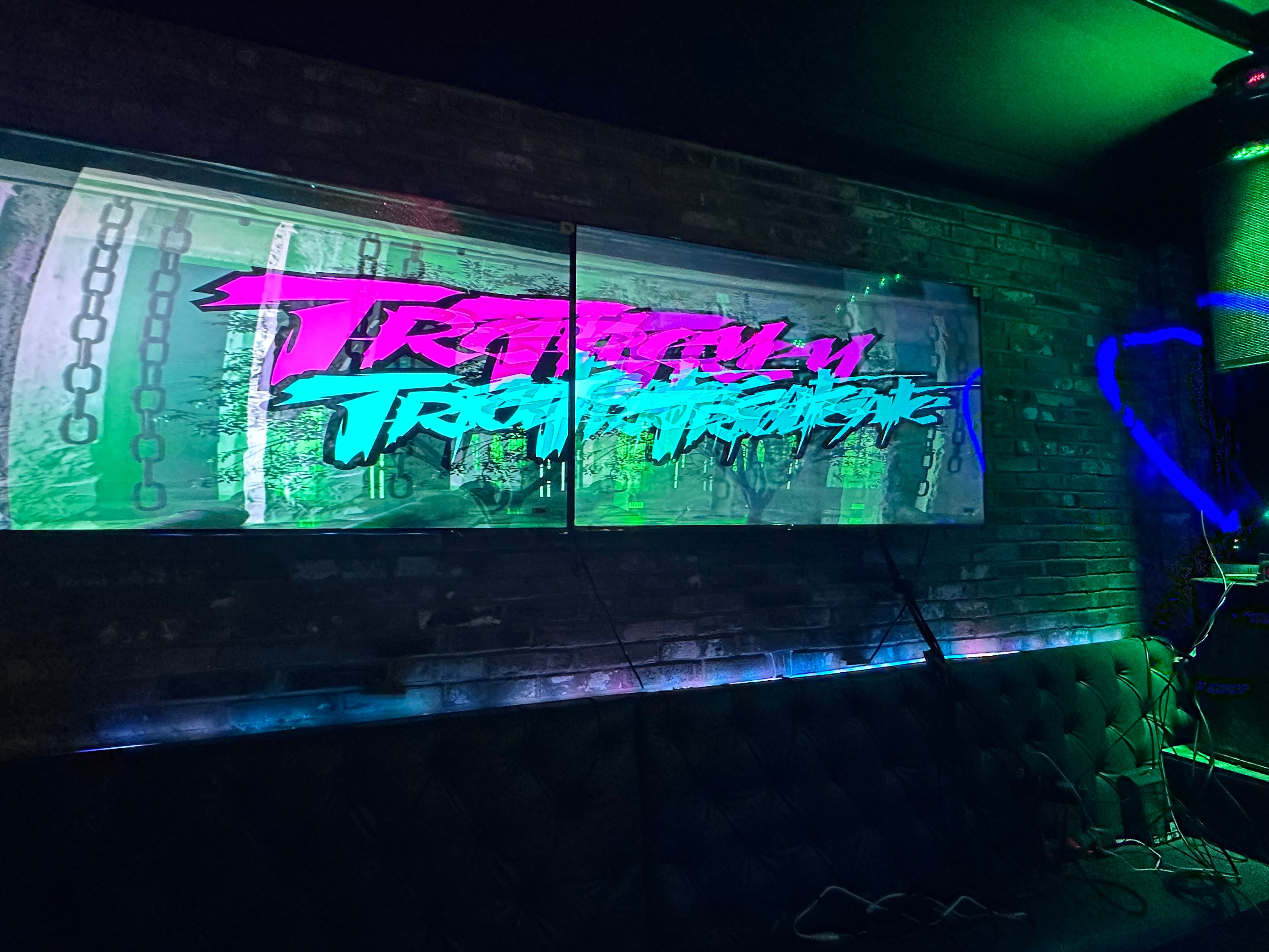 Neon sign with colorful text on a brick wall, illuminated by green and blue lights, with a dark seating area below.