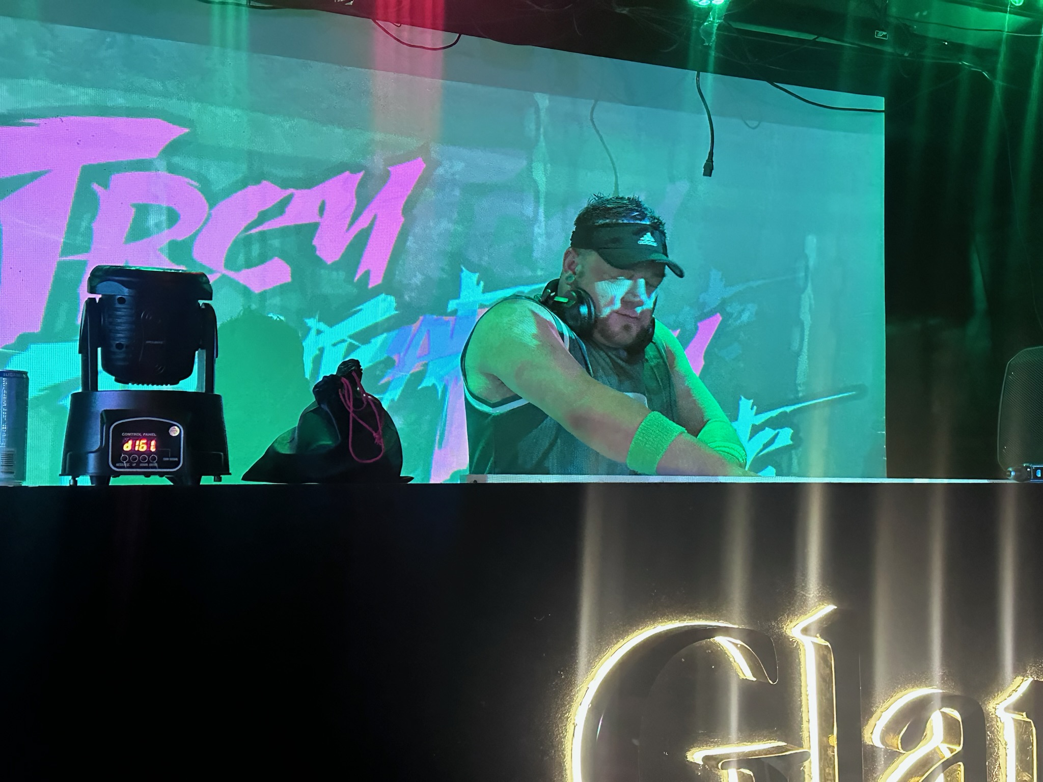 DJ performing on stage with colorful background and lighting effects, wearing headphones and a cap.