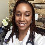 Smiling woman with dreadlocks wearing a white coat and stethoscope, standing in front of a stone wall.
