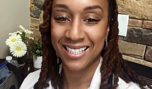 Smiling woman with dreadlocks wearing a white coat and stethoscope, standing in front of a stone wall.