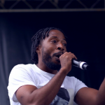 Person speaking into a microphone with a dark background, gesturing with one hand, wearing a white shirt.