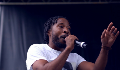Person speaking into a microphone with a dark background, gesturing with one hand, wearing a white shirt.