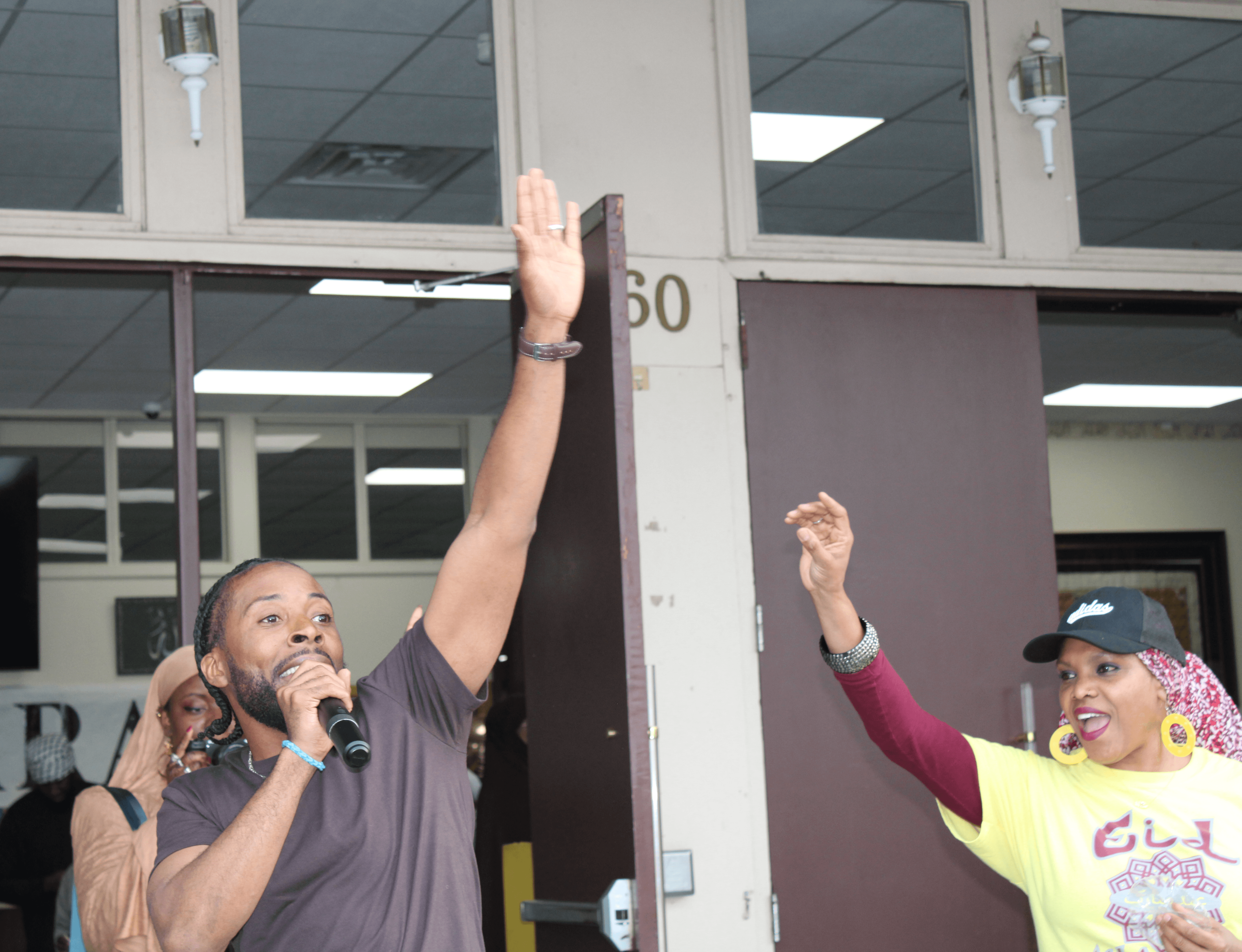 Two people raising their hands, one holding a microphone, inside a building with glass windows and a door numbered 60.