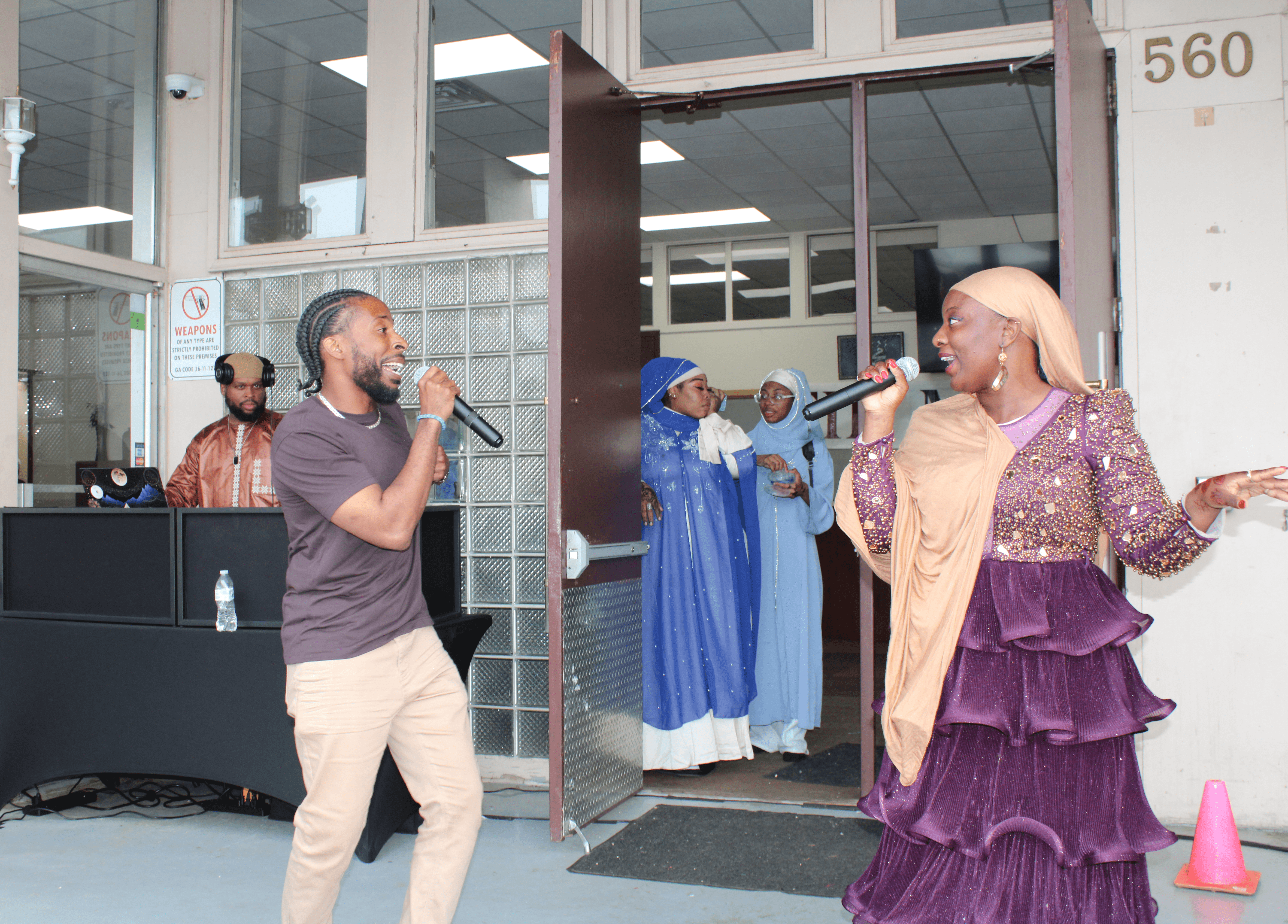 Two people singing into microphones outside a building, with a group of women in blue dresses inside. One woman wears a purple dress and headscarf.