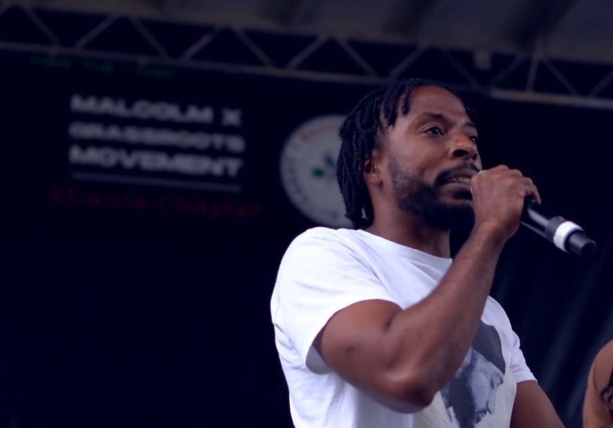 Man speaking into a microphone on stage with a dark background and a banner, wearing a white T-shirt.