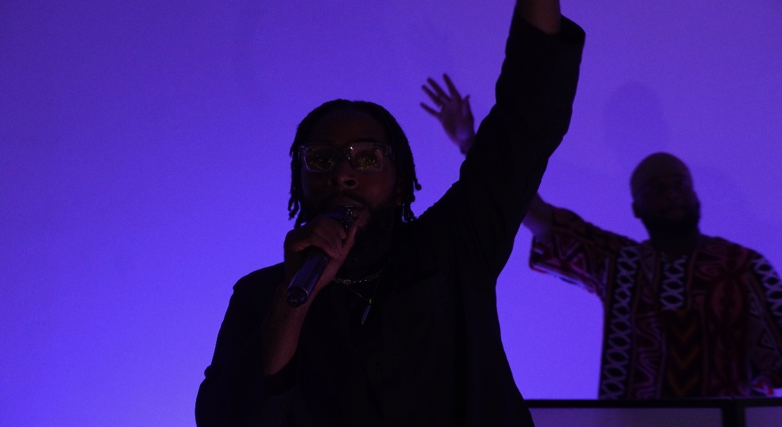 Person holding microphone with one arm raised, silhouette against purple background, another person in background.