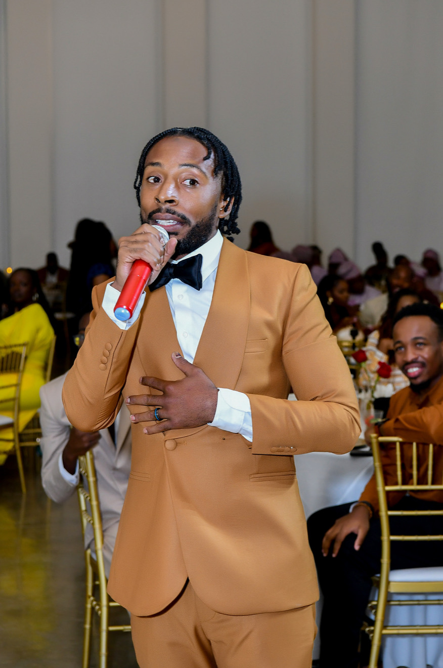Man in a tan suit and black bow tie speaking into a red microphone at a formal event, with seated guests in background.