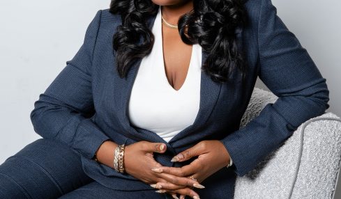 Woman with long black hair, wearing a dark blazer and white top, sitting on a light gray chair against a plain background.