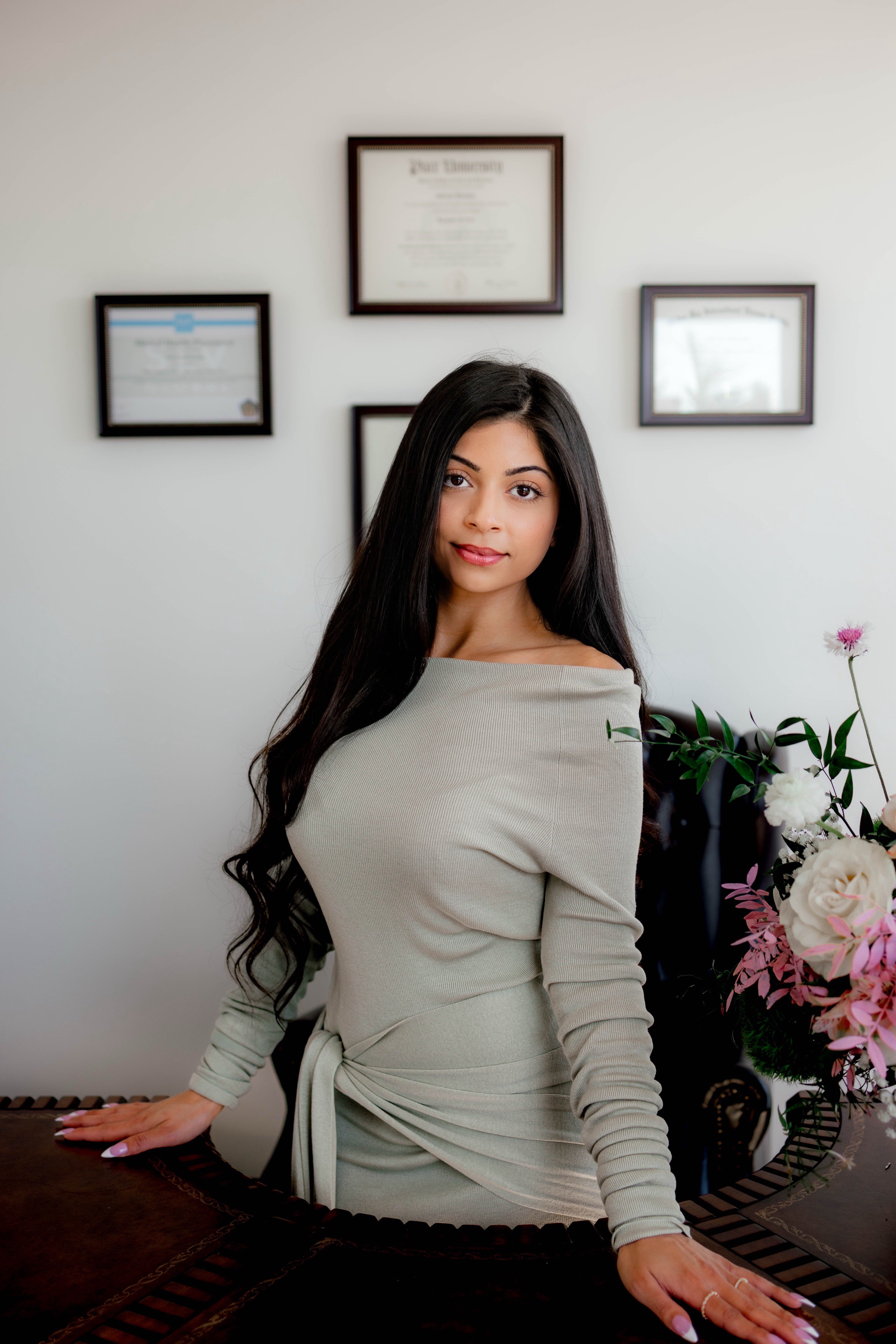 Young woman with long dark hair in a light-colored dress, standing in front of a wall with framed certificates, next to a flower arrangement.