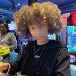 Young person with curly hair in a black jacket in an indoor arcade with colorful lights.
