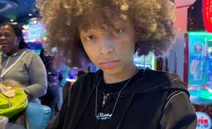 Young person with curly hair in a black jacket in an indoor arcade with colorful lights.