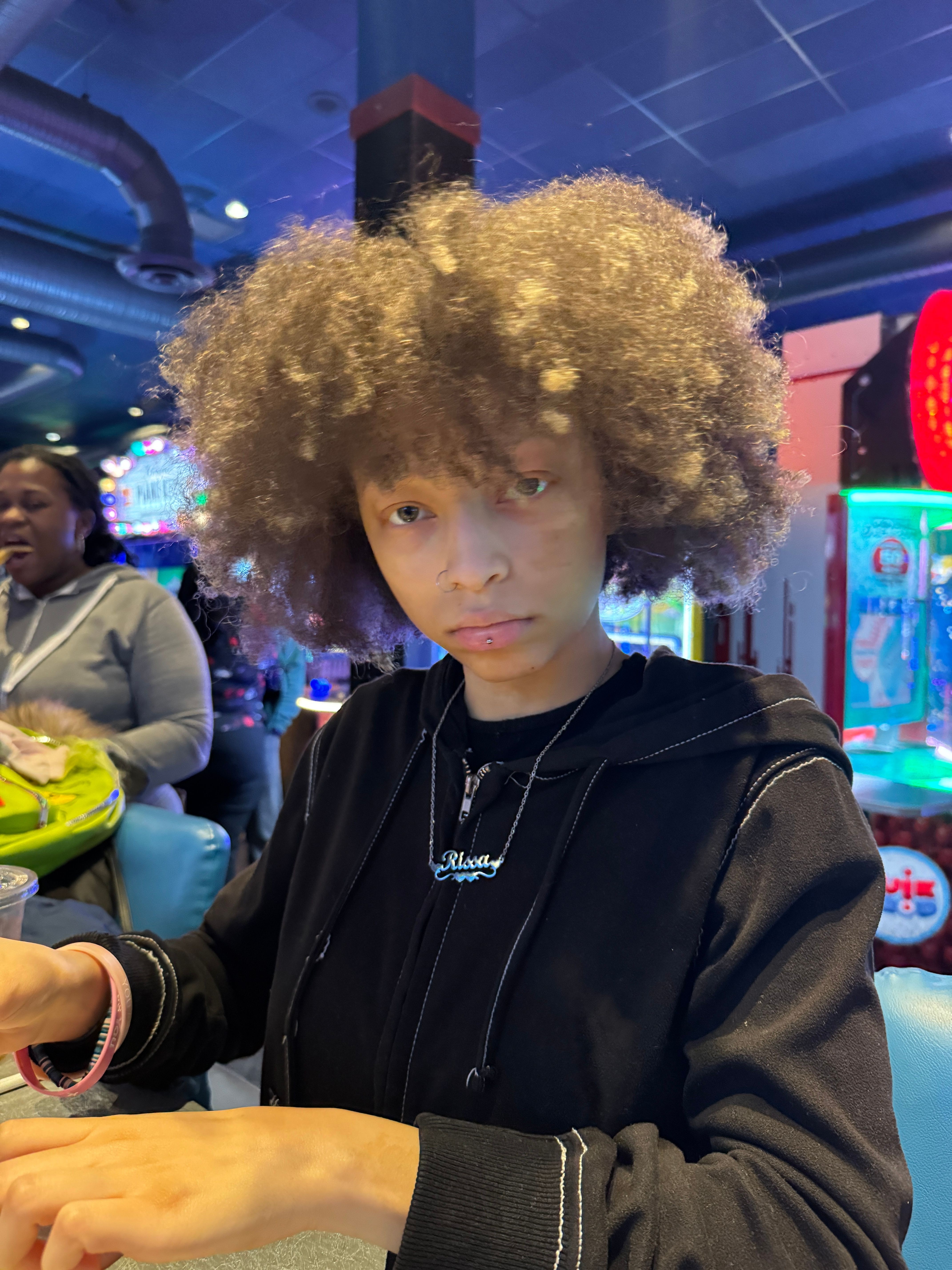 Young person with curly hair in a black jacket in an indoor arcade with colorful lights.