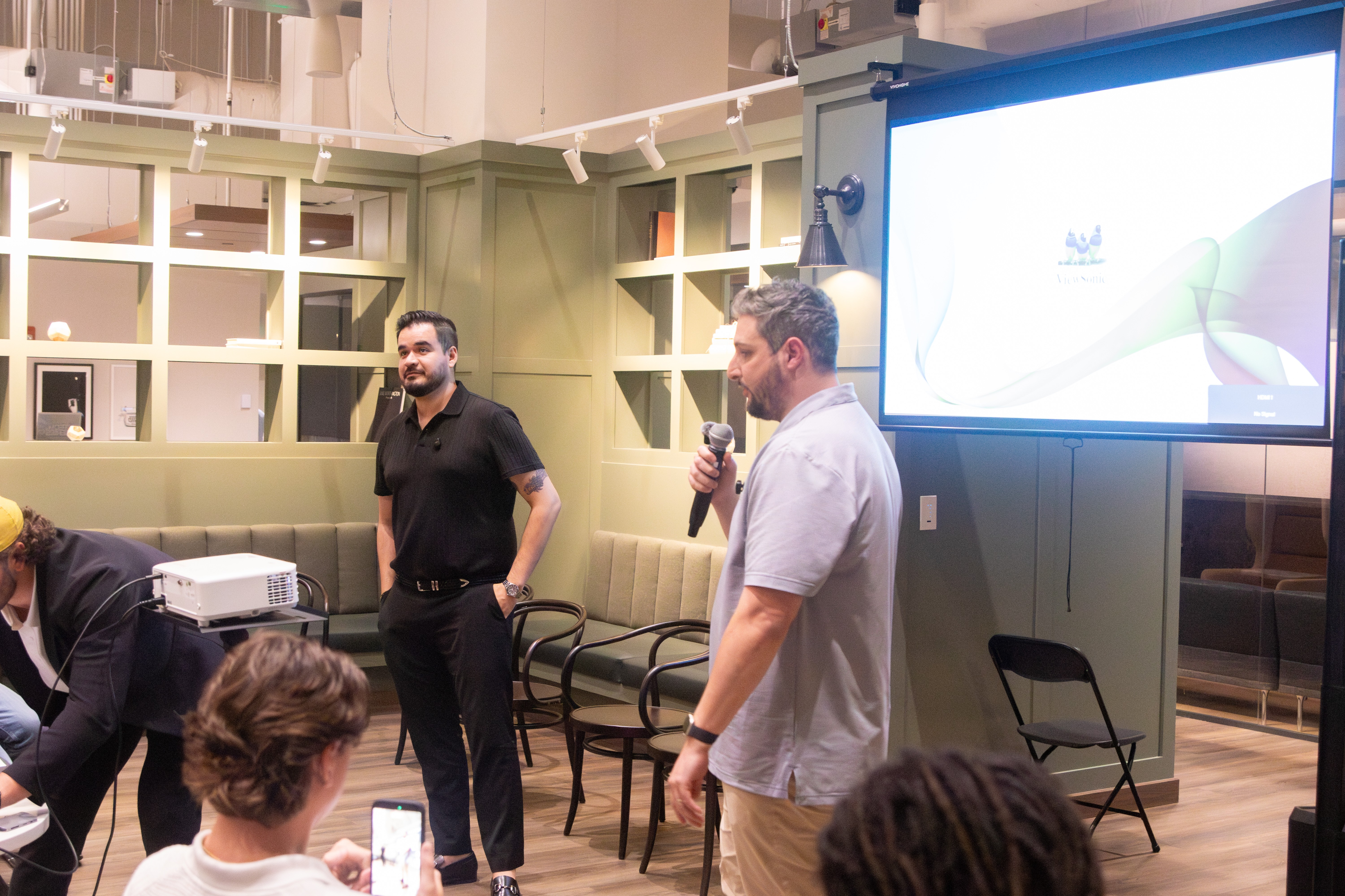 Man speaking into microphone in a room with people, chairs, and a large screen, with a man smiling nearby.