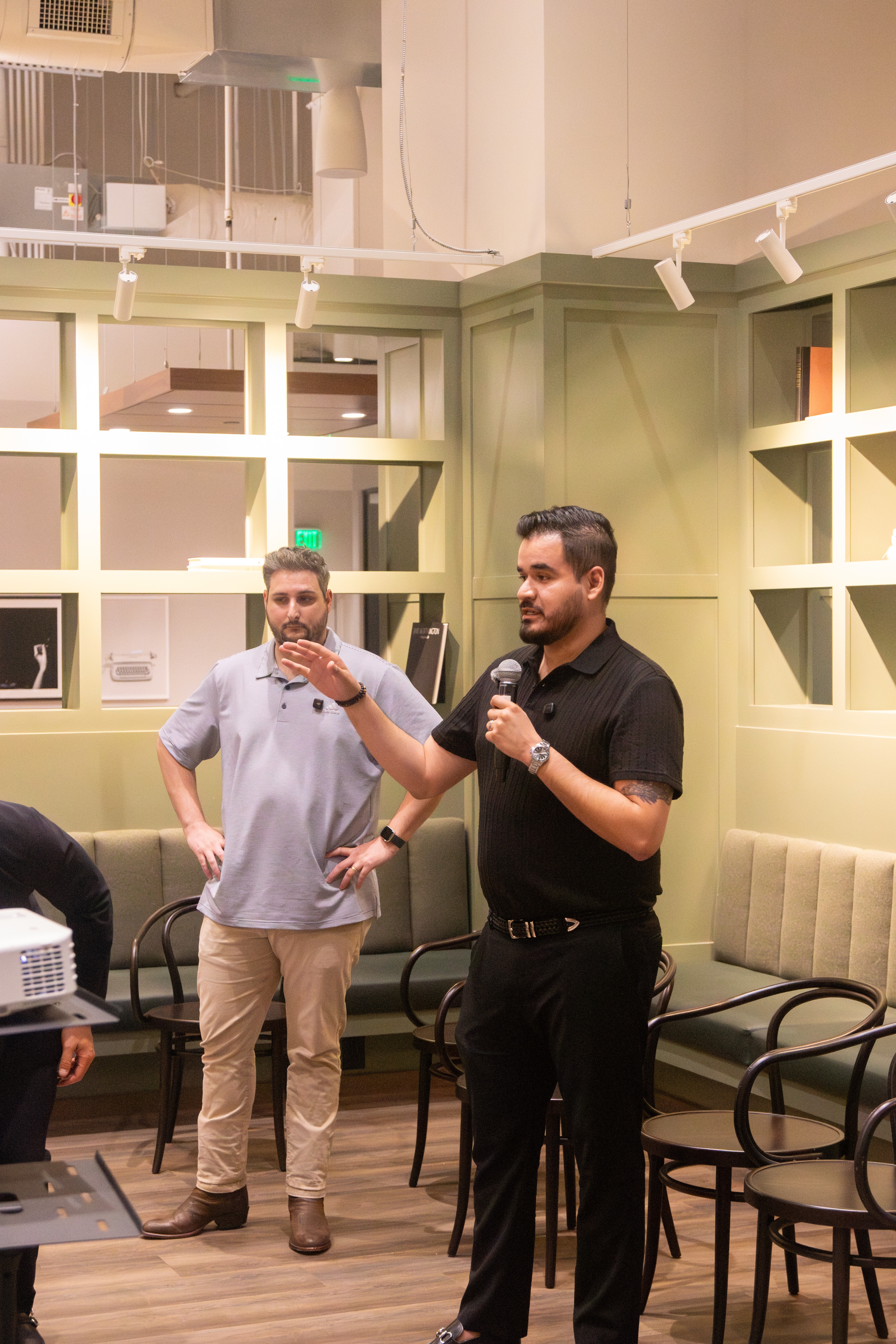Two men stand in a room with green shelves and chairs, one speaking into a microphone, the other listening.