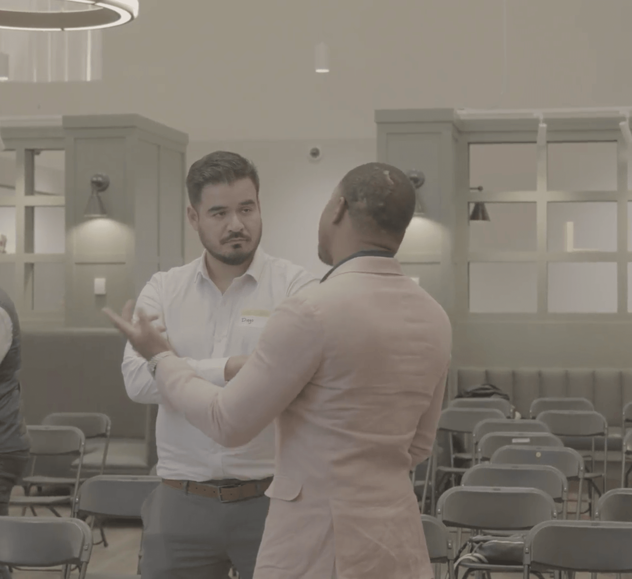 Two men are engaged in conversation in a room with chairs and cabinets, one facing the camera and the other with back turned.