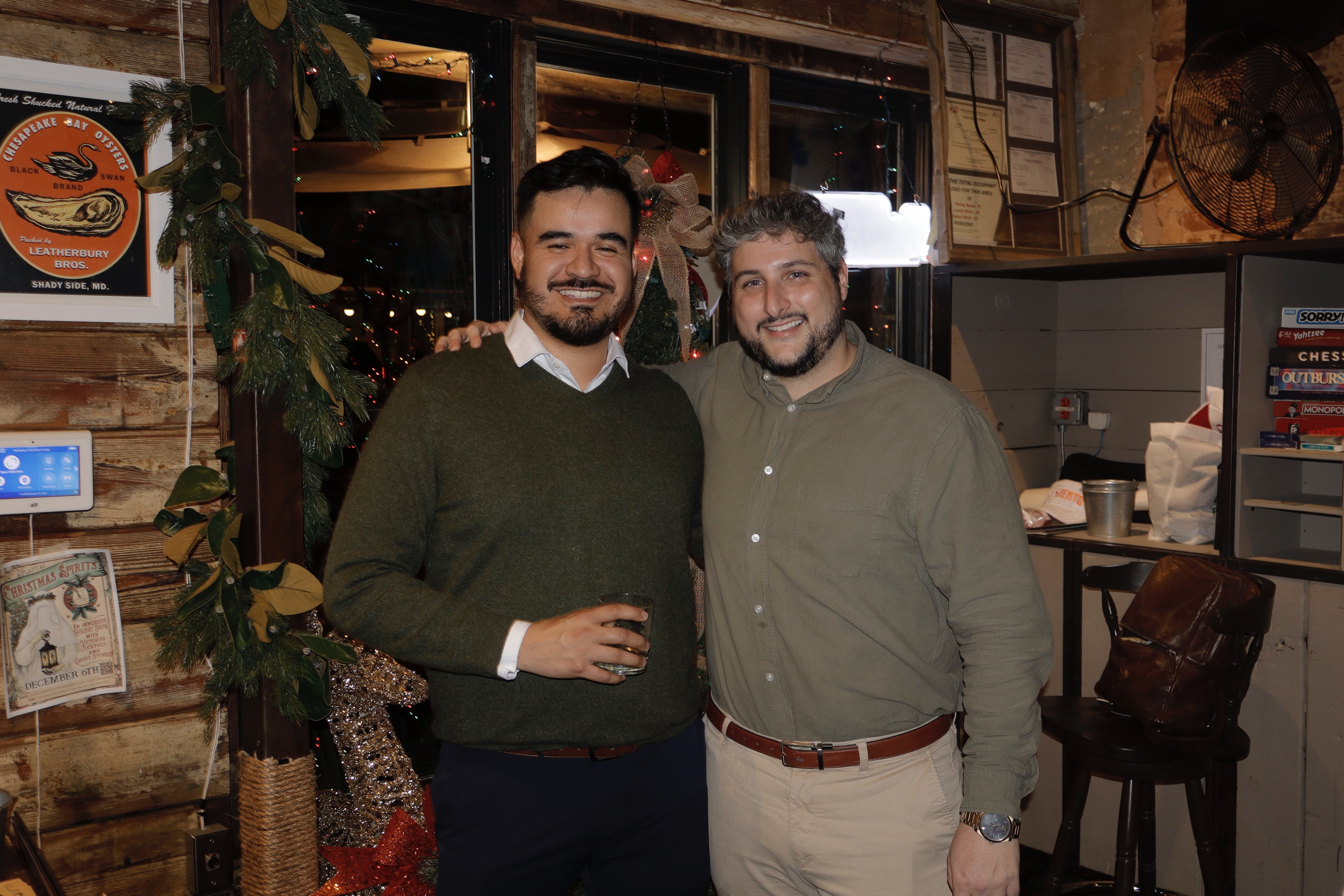 Two men smiling and standing close indoors, one holding a drink, with wooden decor and plants in background.