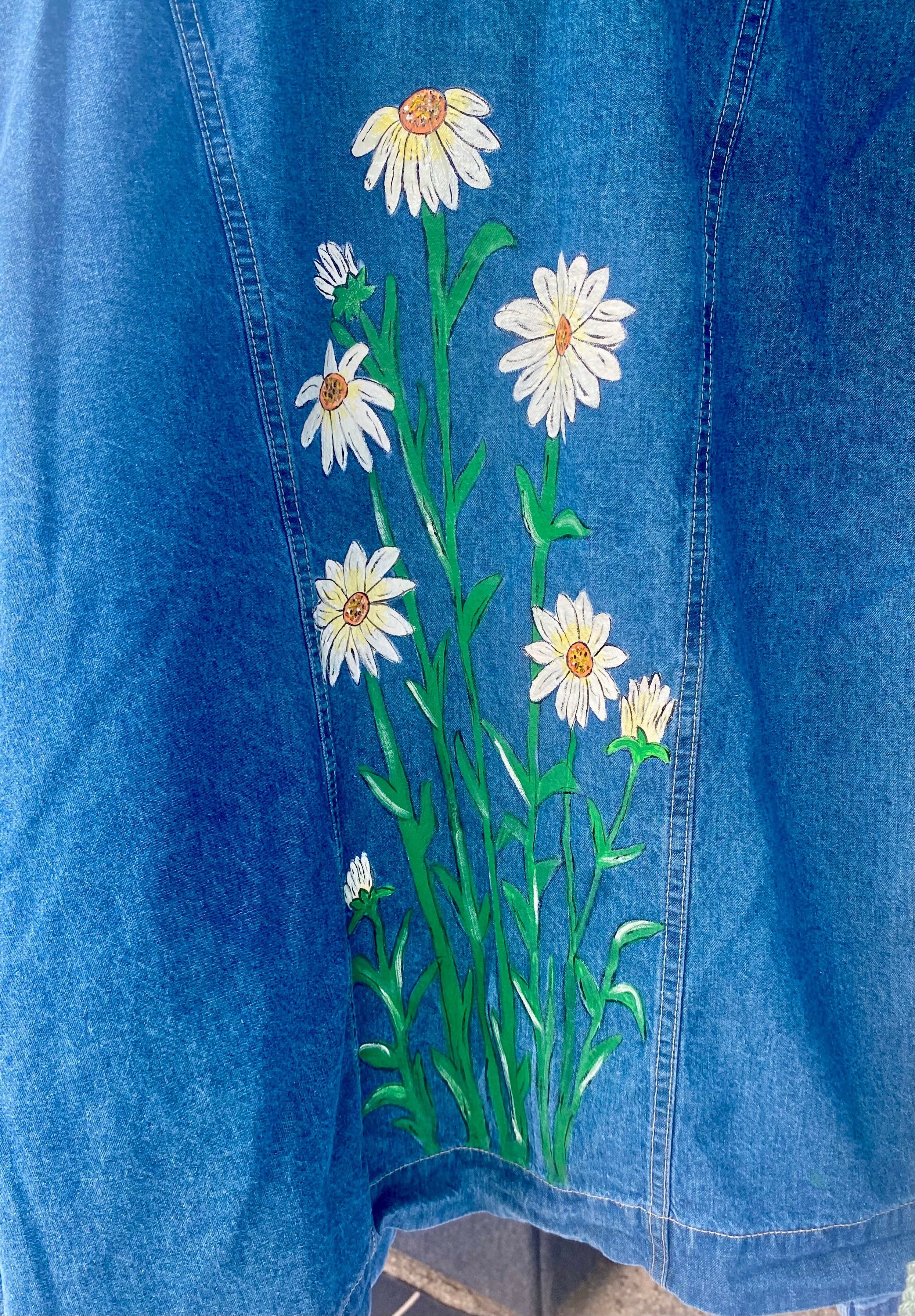 Blue denim fabric with embroidered white daisies and green stems, floral design on the fabric.