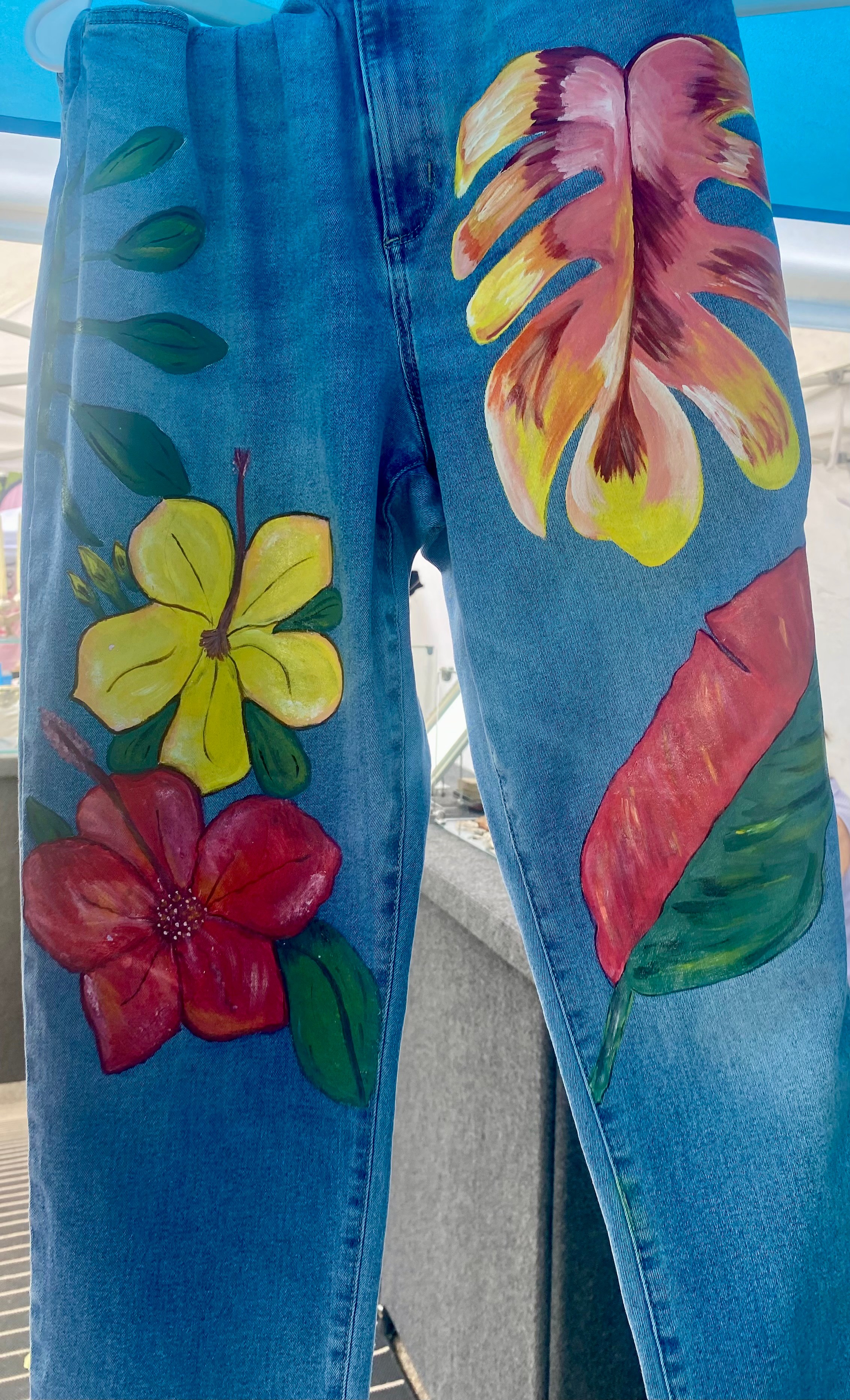 Blue jeans with colorful painted flowers and leaves, including yellow, red, and pink flowers, and green leaves.