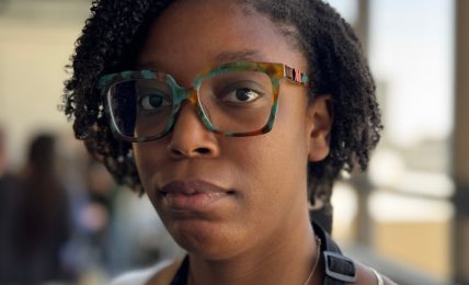 Young woman with glasses and natural hair, indoors near large windows, looking at the camera.