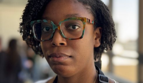 Young woman with glasses and natural hair, indoors near large windows, looking at the camera.