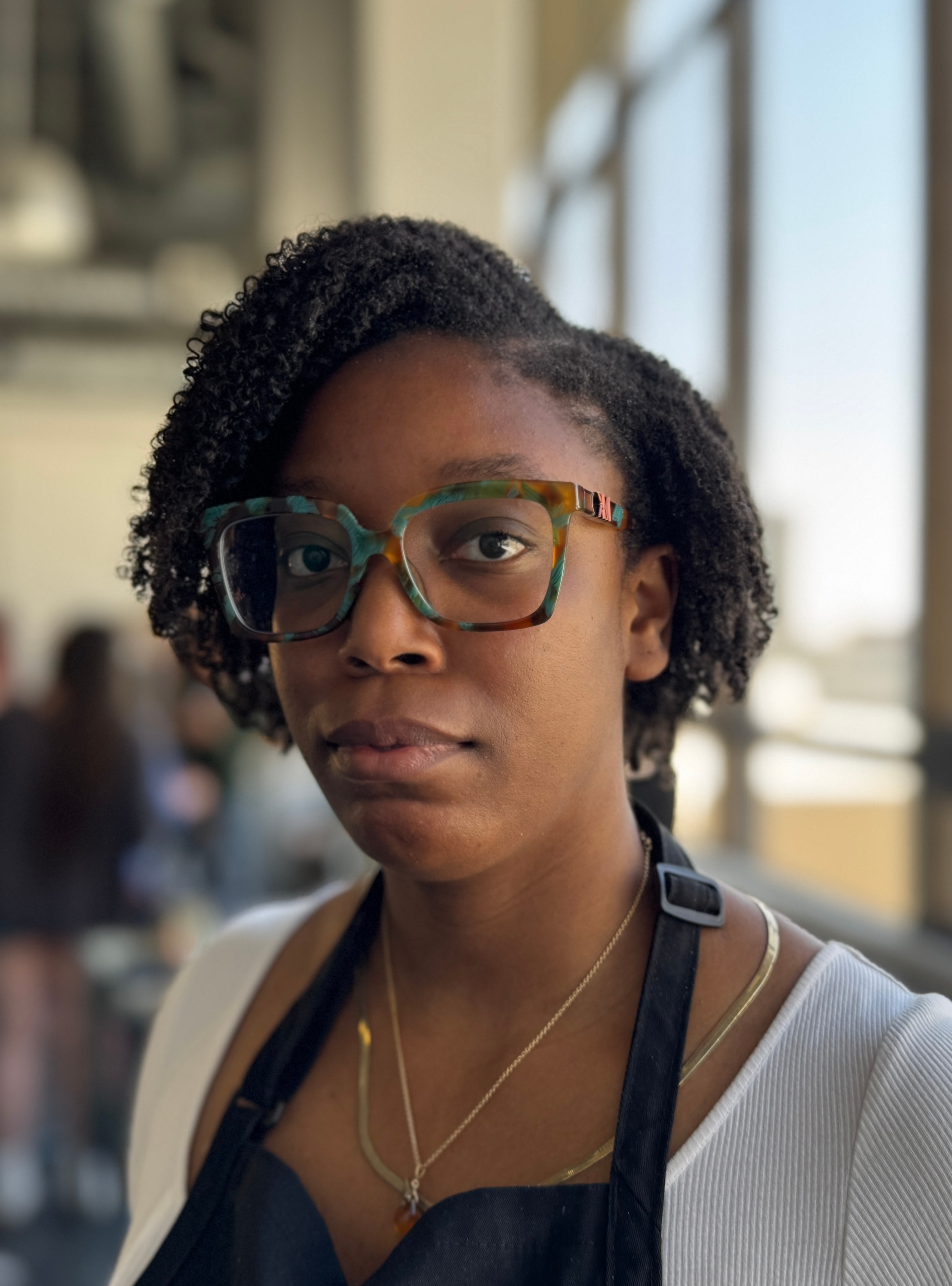 Young woman with glasses and natural hair, indoors near large windows, looking at the camera.