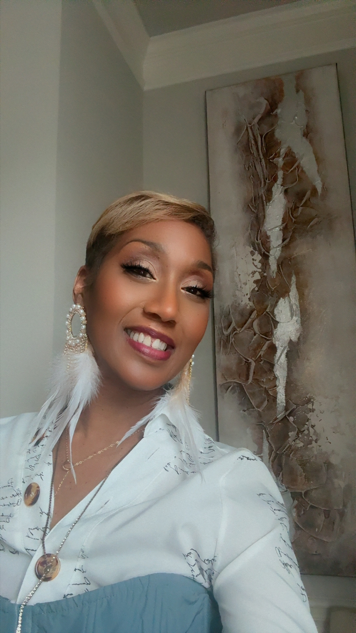 Smiling woman with short hair, wearing earrings and a white top, standing indoors near abstract wall art.