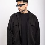 Man with dark hair and beard wearing sunglasses, black jacket, and black shirt, looking to the side against a white background.