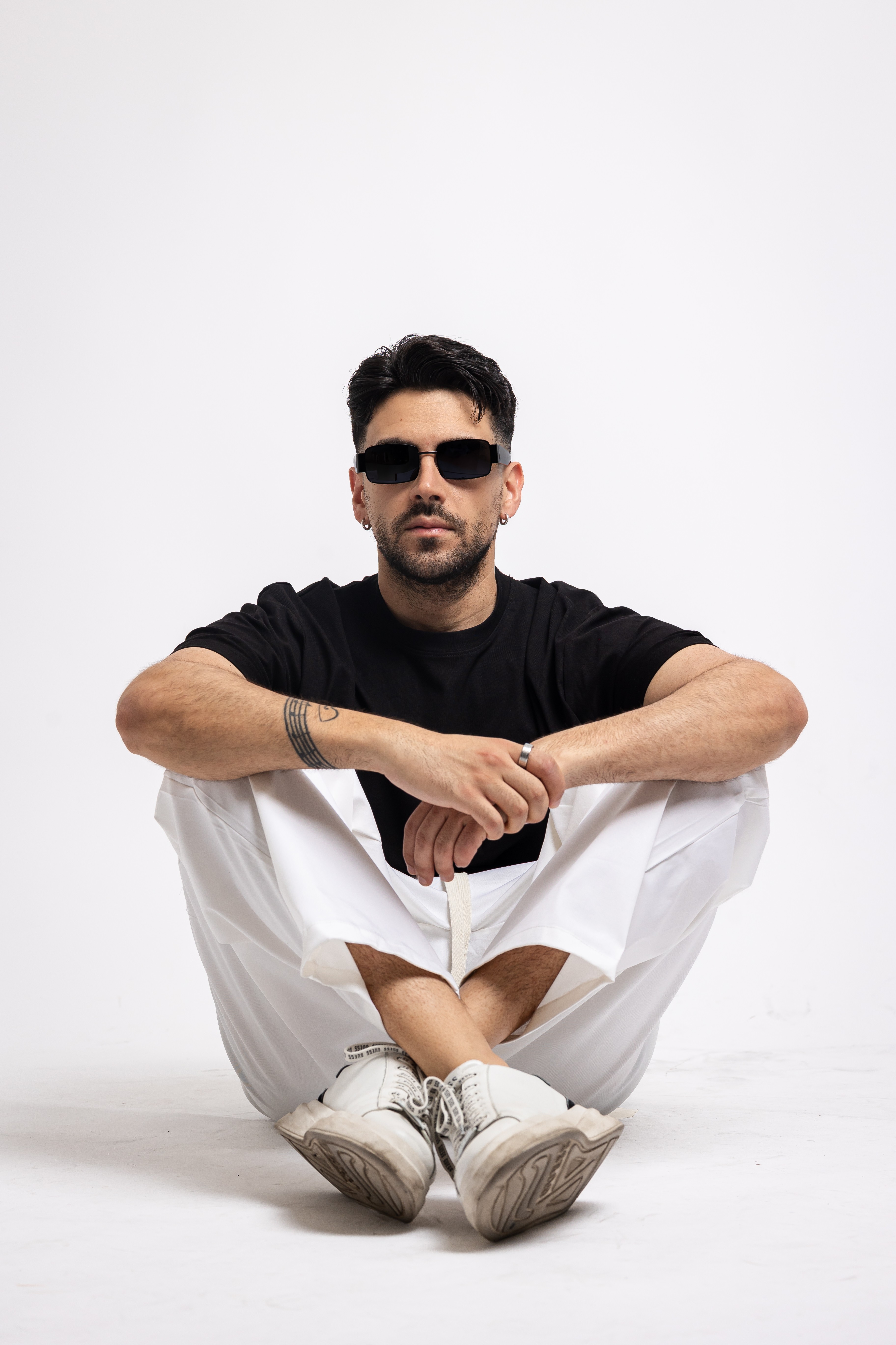 Man sitting cross-legged on the floor, wearing sunglasses, black t-shirt, white pants, and sneakers, against a plain white background.
