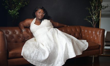 Woman in a white dress sitting on a brown leather sofa with plants in the background.