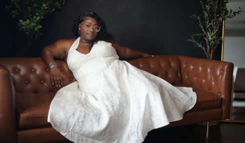 Woman in a white dress sitting on a brown leather sofa with plants in the background.