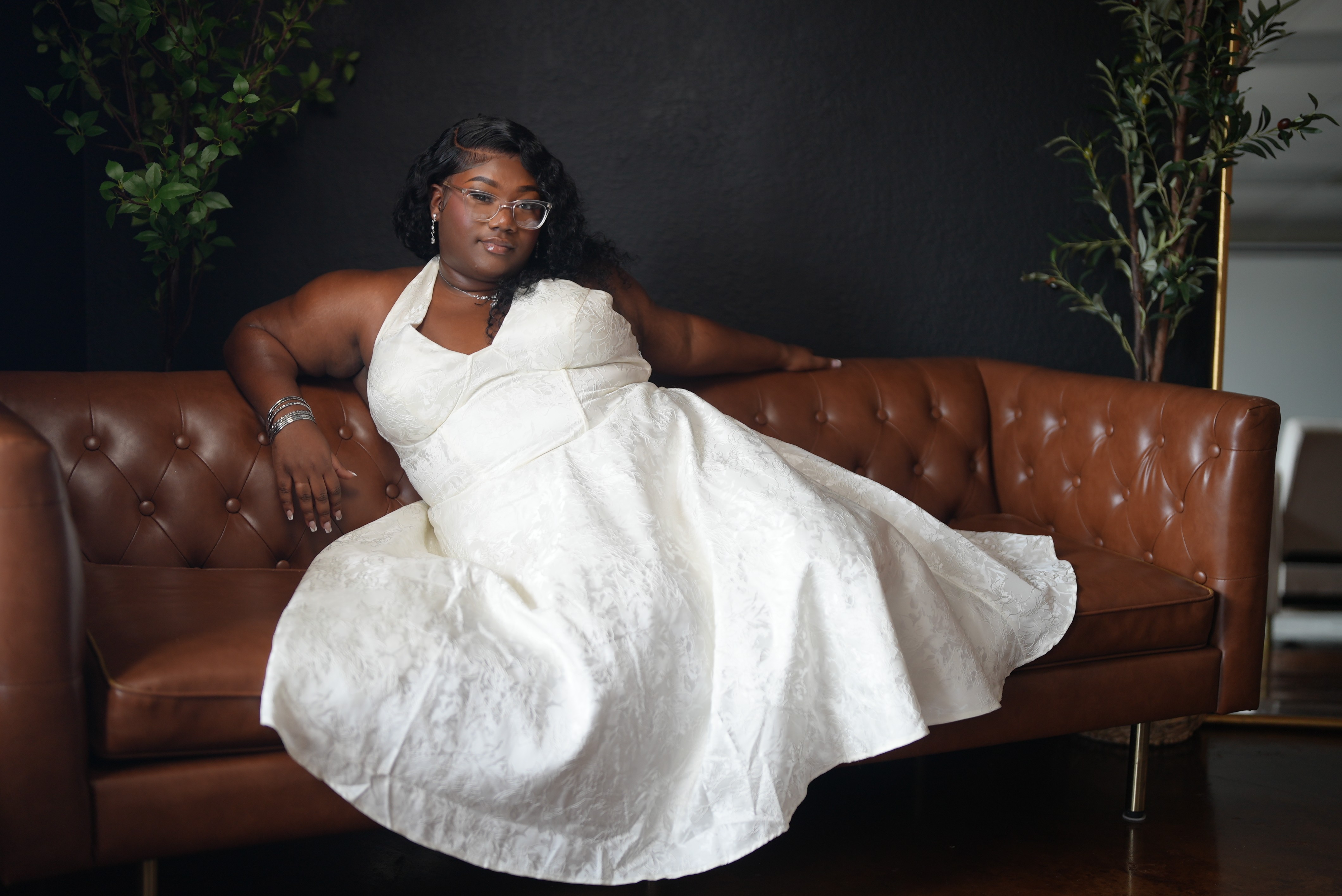 Woman in a white dress sitting on a brown leather sofa with plants in the background.