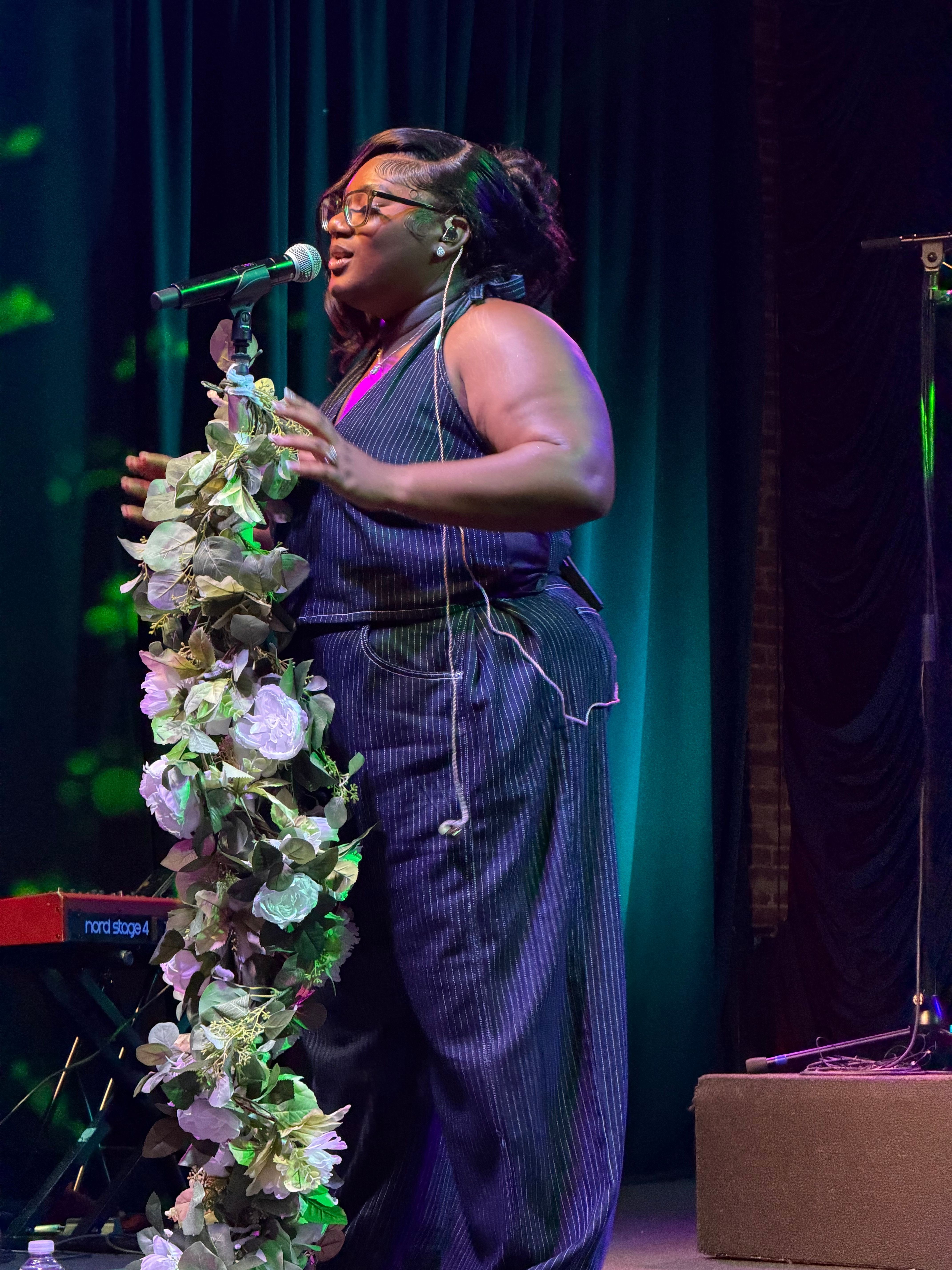 Person with long hair and glasses speaking into a microphone on stage, decorated with flowers, in front of a dark curtain.