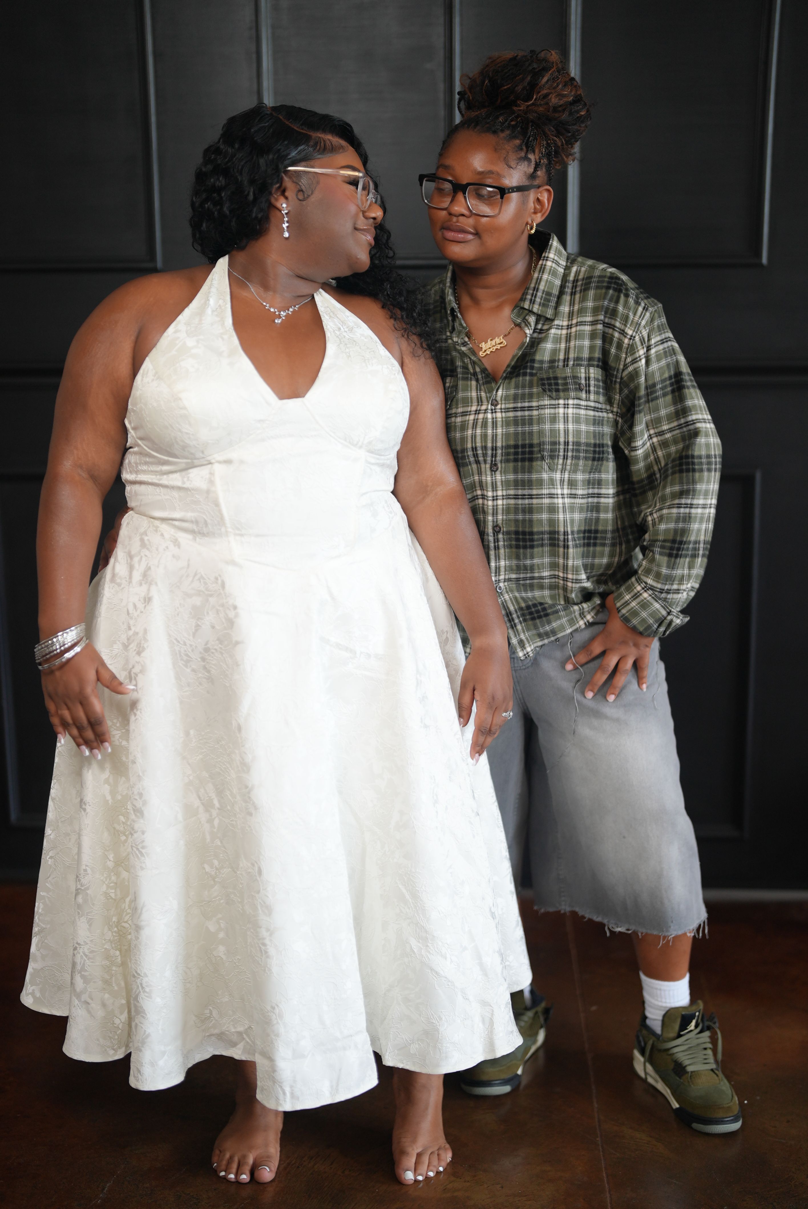 Two women standing together, one in a white dress and the other in a plaid shirt and gray skirt, in front of a dark background.