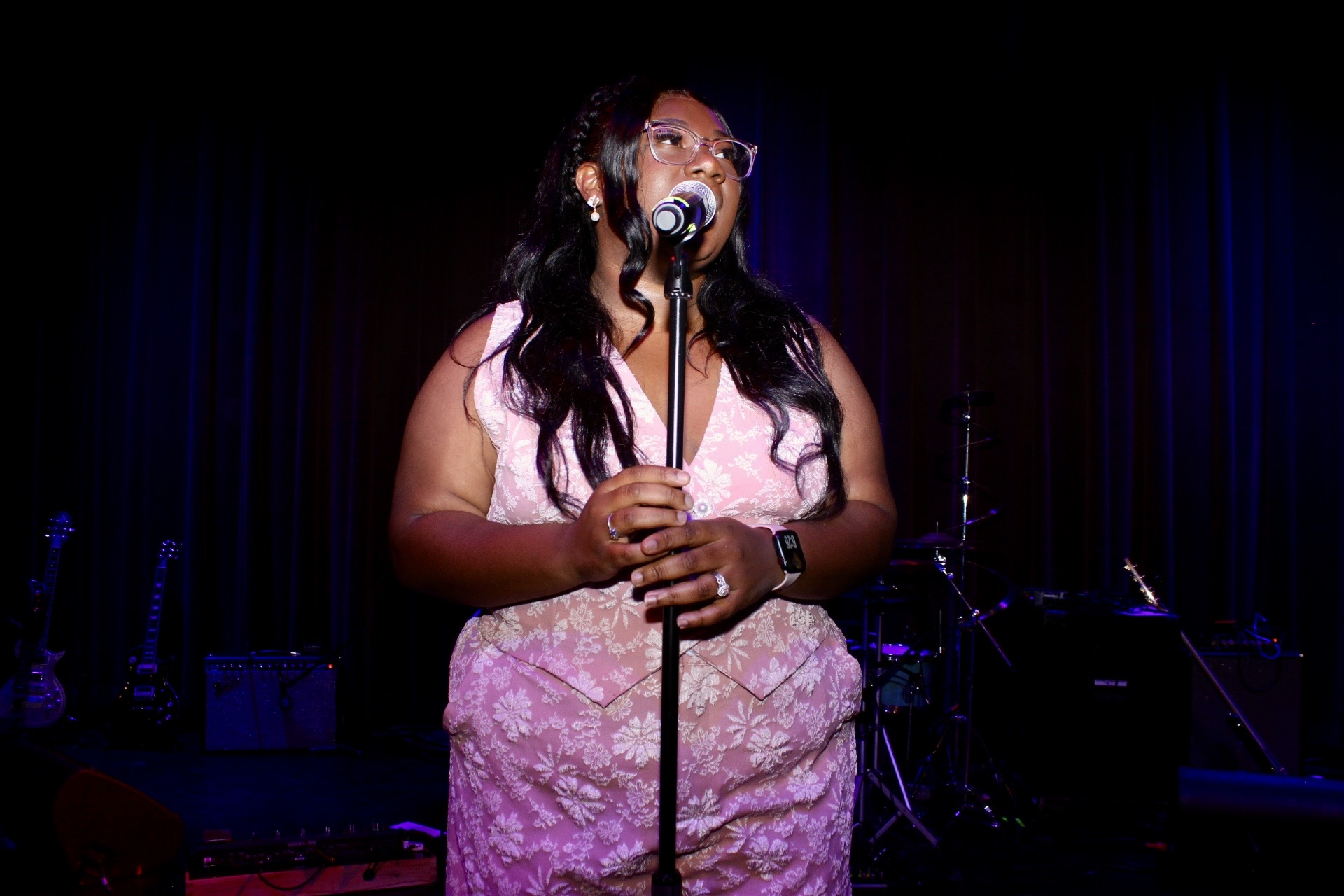 A woman in a pink dress stands on stage holding a microphone, with dark curtains in the background.