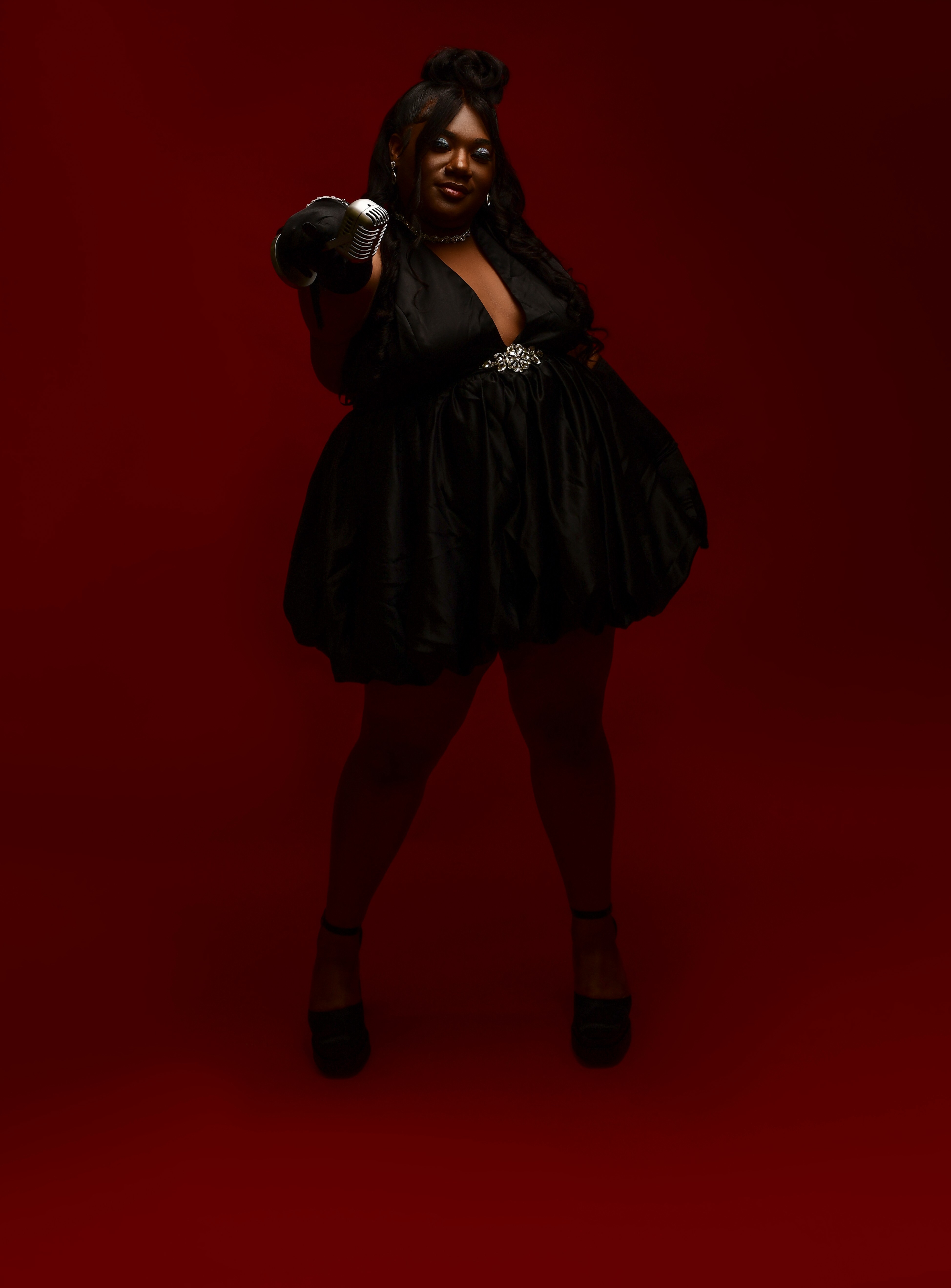 Person in black dress pointing forward against a red background, with dark hair and glasses.