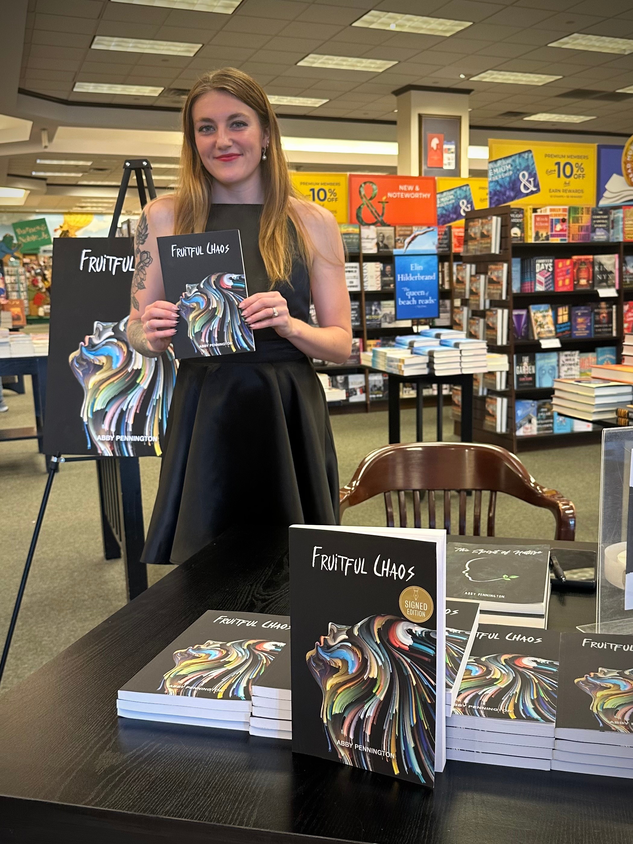 Young woman with long hair holding a book in a bookstore, standing behind a table with multiple copies of the book, bookshelves in background.