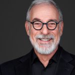 Portrait of an older man with glasses, gray hair, and a beard, smiling against a dark background.