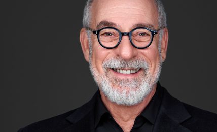 Portrait of an older man with glasses, gray hair, and a beard, smiling against a dark background.