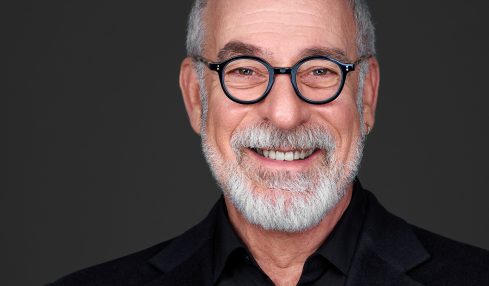 Portrait of an older man with glasses, gray hair, and a beard, smiling against a dark background.