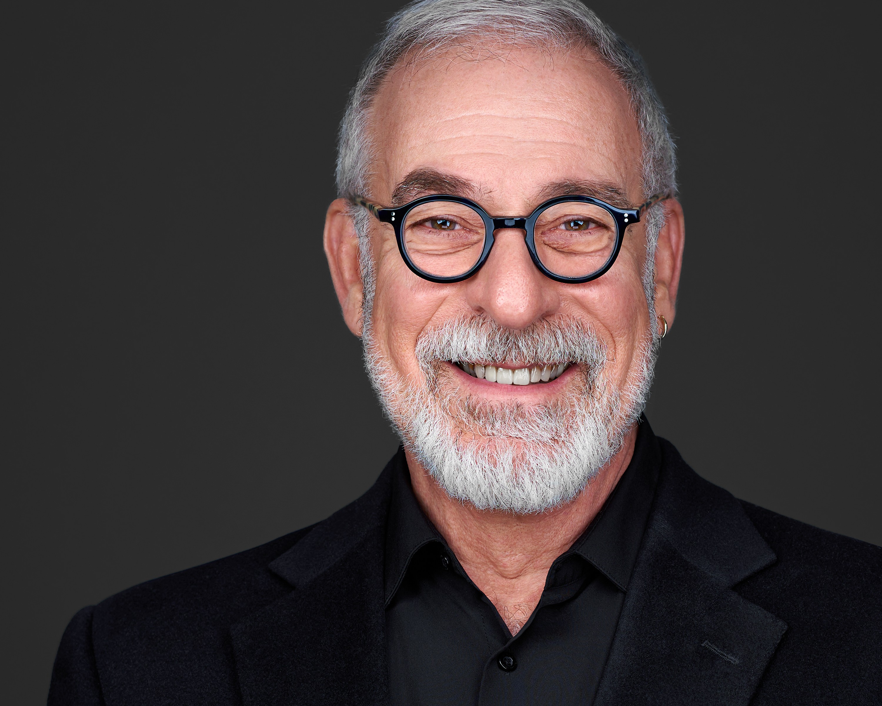 Portrait of an older man with glasses, gray hair, and a beard, smiling against a dark background.