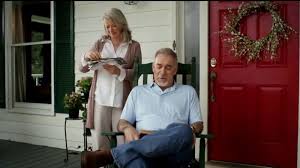 An elderly woman stands reading a book or magazine, while an elderly man sits on a porch chair with arms crossed.